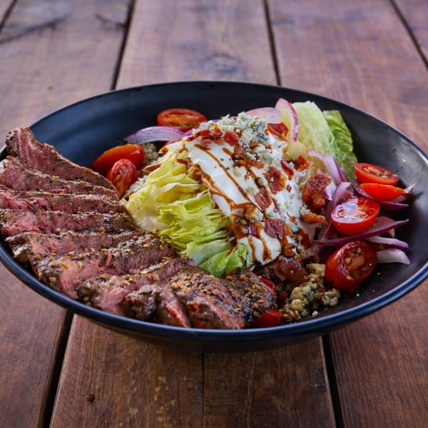 Steak and wedge salad with blue cheese, bacon, tomatoes, and red onion in a black bowl.