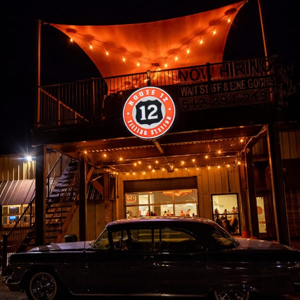 A vintage car parked outside Route 12 Filling Station at night, with string lights and a glowing sign.