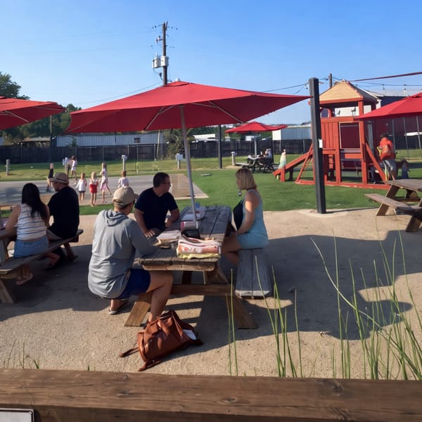 Patrons sit at picnic tables under red umbrellas, with a playground and children in the background.
