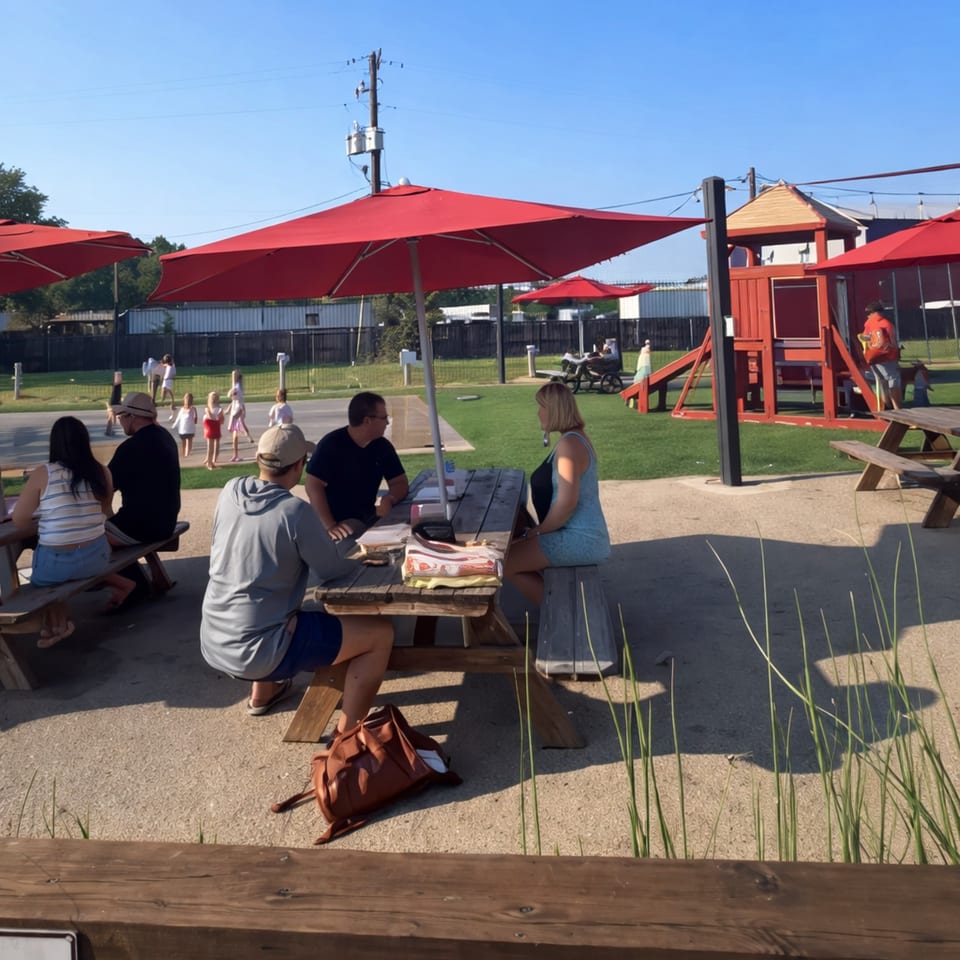 Patrons sit at picnic tables under red umbrellas, with a playground and children in the background.