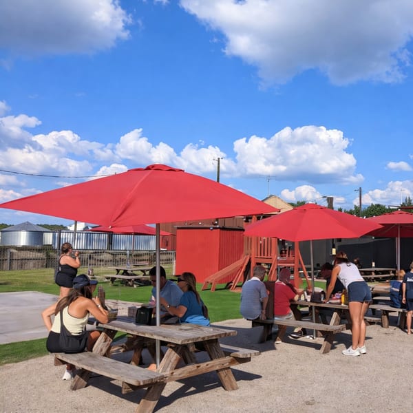 People enjoying outdoor dining at picnic tables under red umbrellas on a sunny day.