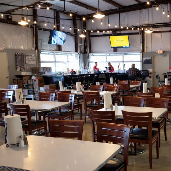 Spacious restaurant interior with white tables, wooden chairs, a bar, and string lights overhead.
