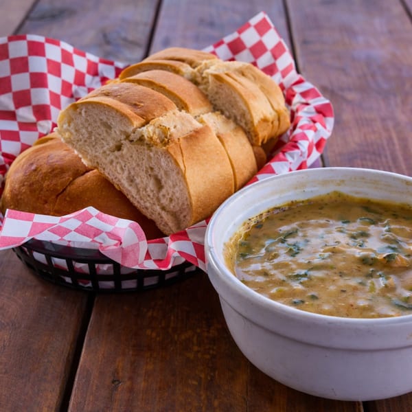 Sliced baguette in a basket with red checkered paper, next to a bowl of creamy soup.