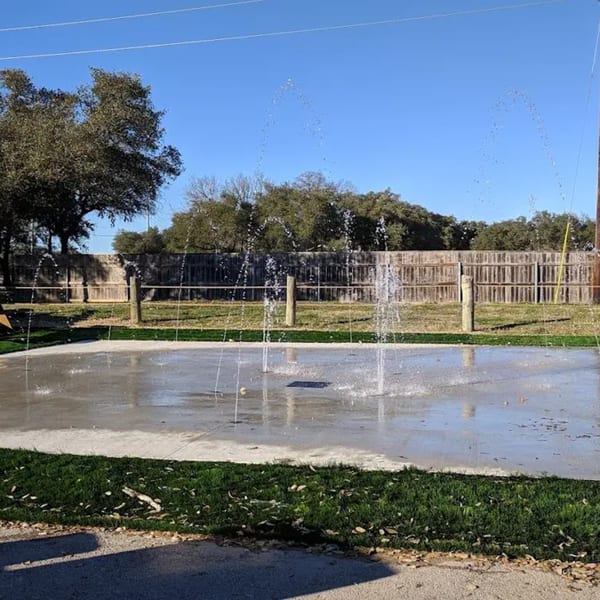 A splash pad with water spraying upwards, surrounded by green grass and a wooden fence.