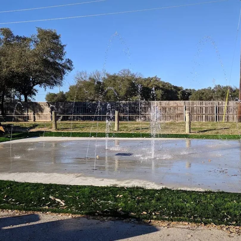 A splash pad with water spraying upwards, surrounded by green grass and a wooden fence.