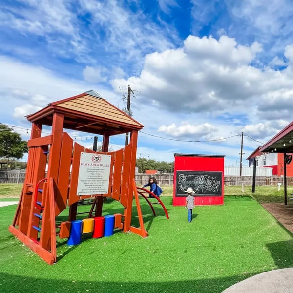 Kids playing on a red wooden playground with a chalkboard wall on green turf under a blue sky.