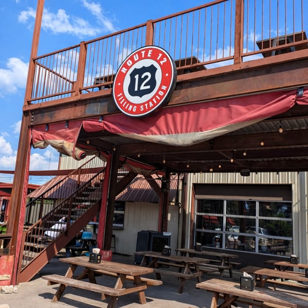 The exterior of Route 12 Filling Station, featuring a red and white sign, picnic tables, and an upper deck.