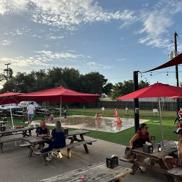 People dine at outdoor picnic tables under red umbrellas, with children playing in a splash pad nearby.